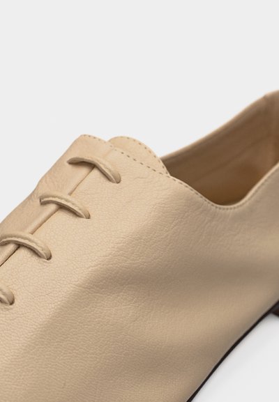 Μπεζ δερμάτινο παπούτσι με λείο υφή και μοναδικό σχεδιασμό κορδονιών σε στυλ θηλιάς στο πάνω μέρος, εμφανιζόμενο σε λευκό φόντο.