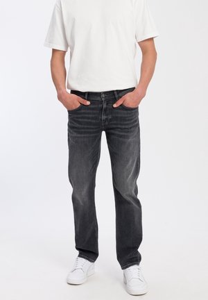 Homme portant un jean noir délavé, des baskets blanches et une chemise blanche à manches courtes, debout les mains dans les poches sur fond blanc.