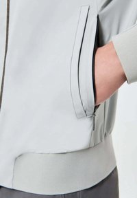 Giacca grigia chiara con una texture liscia, dotata di una tasca con cerniera sul lato e di una vita a coste. Design minimalista e linee pulite.