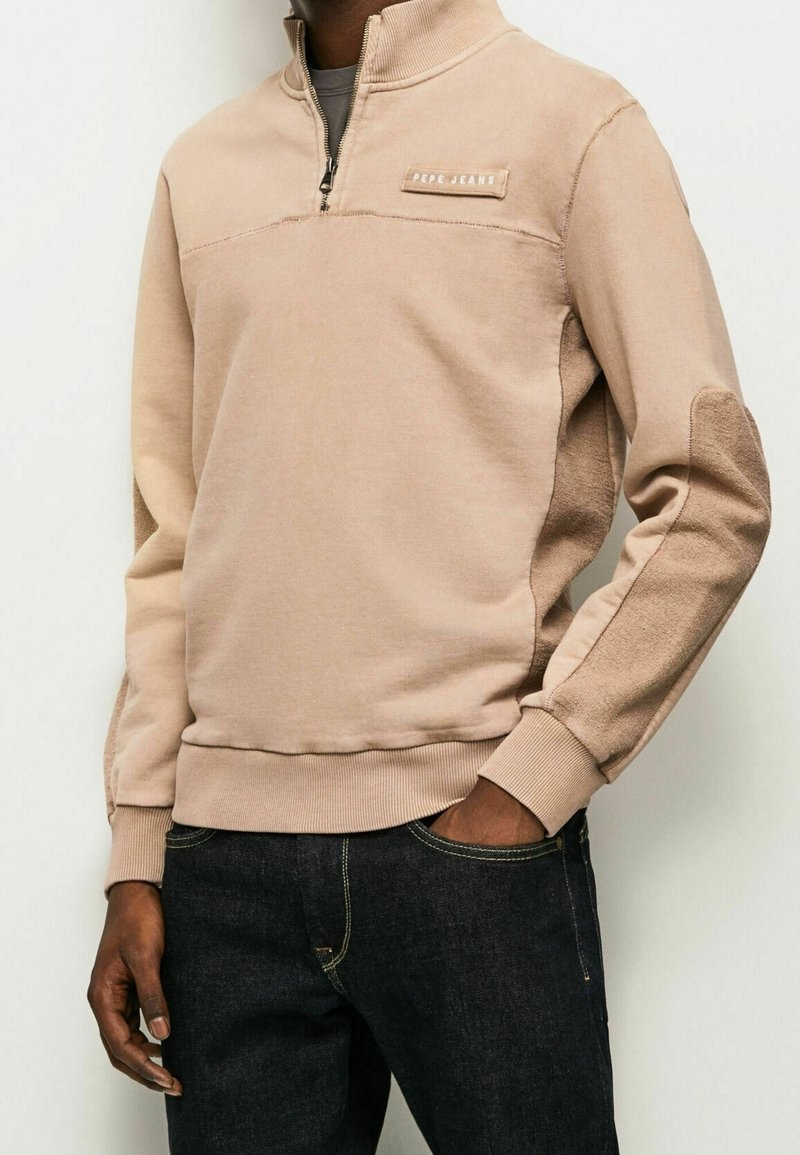 Sudadera beige con cremallera hasta el cuarto, panel texturizado, puños de canalé y un pequeño parche de logo en el pecho. Combinada con jeans oscuros.