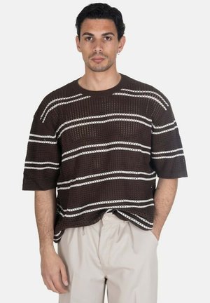 Uomo che indossa un maglione corto a maniche corte marrone scuro a maglia con strisce orizzontali bianche e pantaloni beige chiaro, in piedi con una mano in tasca.