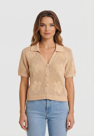 Mujer con cárdigan de punto beige de manga corta con estampados florales y botones, combinado con vaqueros de cintura alta de color azul claro, de pie sobre un fondo neutro.