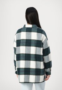 Koszula w kratę oversize w ciemnozielonym i białym kolorze, z klasycznym kołnierzykiem, długimi rękawami oraz guzikami na mankietach. Miękka w dotyku.