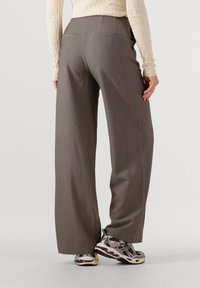 Osoba nosząca szare spodnie o szerokich nogawkach, beżową teksturowaną bluzkę z długim rękawem oraz białe i fioletowe sportowe buty, stojąca na tle jednolitego tła.