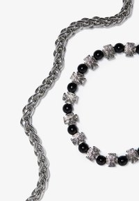 Sølvmetallkjede med flettet design ved siden av et sirkulært perlekjede som har svarte perler og sølvfargede korsformede detaljer.