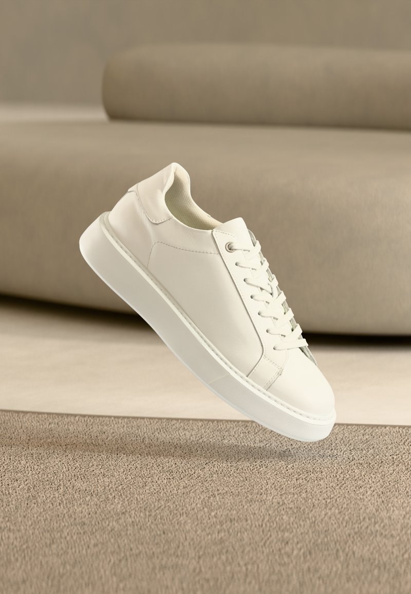 Λευκό δερμάτινο αθλητικό παπούτσι με κορδόνια που αιωρείται πάνω από μπεζ χαλί και καναπέ σε δωμάτιο με ουδέτερους τόνους.