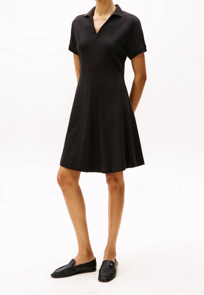 Robe polo noire à manches courtes, col en V, corsage ajusté qui s’évase en jupe. Associée à des chaussures noires sans lacets.