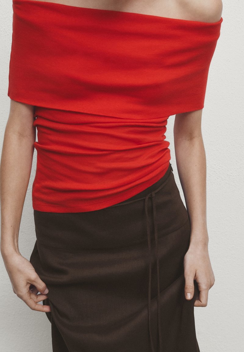 Rotes schulterfreies Top mit geraffter Textur, kombiniert mit einem braunen Rock, der ein Zugbanddetail an der Taille hat. Glatte Stoffoberfläche.
