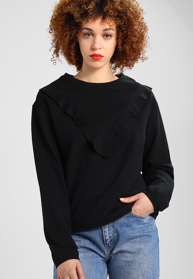 Fekete, hosszú ujjú pulóver, fodros V-nyakkivágással és puha pamut anyagból, világoskék farmernadrággal párosítva.