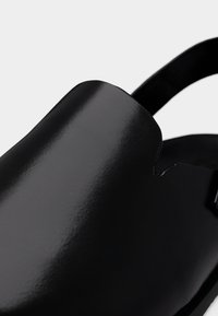 Μαύρα σανδάλια από γυαλιστερό υλικό, με λεία επιφάνεια, ανοιχτή πλάτη και σχέδιο με κοψίματα στο επάνω μέρος. Περιλαμβάνει ρυθμιζόμενο λουράκι.