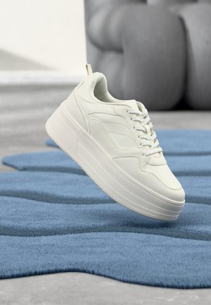White leather sneaker with a thick platform sole, textured detailing, and traditional lacing, positioned above a blue textured rug.