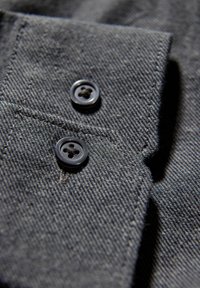 Donkergrijze stof close-up met twee zwarte knopen met vier gaten en zichtbare steken, die de textuur en het weefpatroon benadrukken.