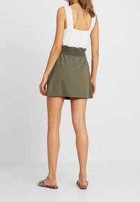 Jupe mini vert olive avec une ceinture froncée, en tissu doux. Associée à un top sans manches, ajusté, de couleur blanche et des sandales plates.