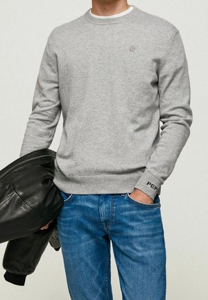 Mand iført lysegrå sweater og blå jeans, holder en sort læderjakke i den ene hånd, den anden hånd i lommen.