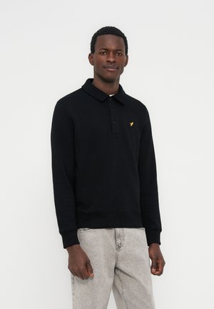 Άνδρας με μαύρο πόλο με μακριά μανίκια και κίτρινο λογότυπο πουλιού στο στήθος, φοράει ανοιχτό γκρι τζιν, στέκεται μπροστά σε απλό ανοιχτό φόντο.