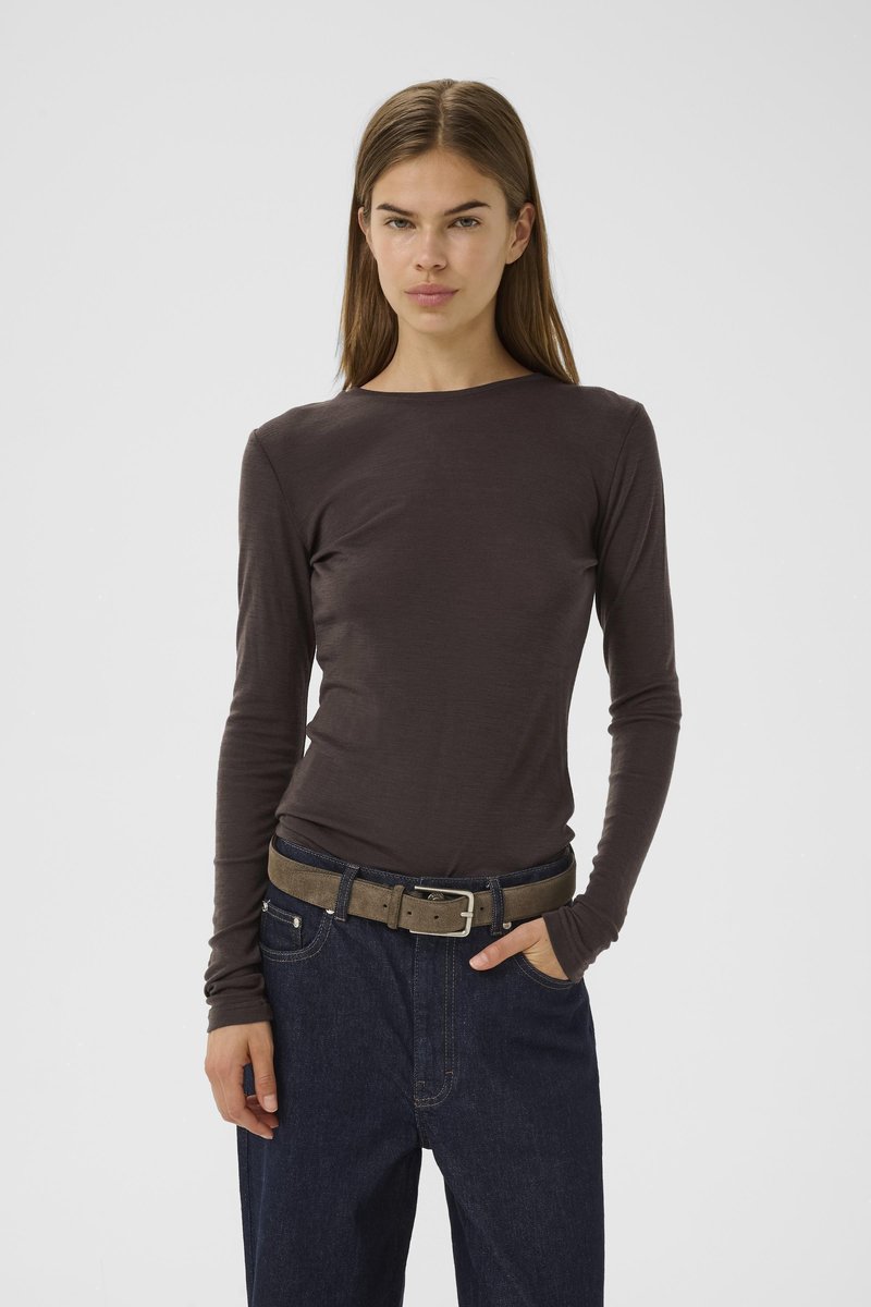 Young person with straight brown hair wearing a dark long-sleeve shirt, blue jeans with a brown belt, one hand in pocket.