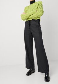 Grön stickad tröja med kabelmönster, matchad med svarta vida byxor med dragsko i midjan. Modellen bär svarta skor.