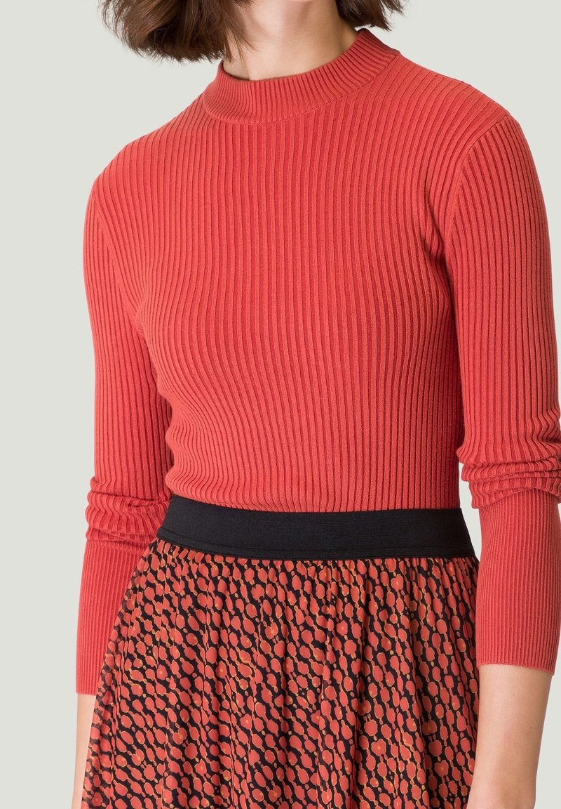 Haut à manches longues côtelé rouge avec un col montant, associé à une taille noire et une jupe à motifs comportant des pois rouges et orange.