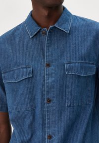 Camicetta in denim a maniche corte con motivo a spina di pesce blu. Presenta un colletto con bottoni, due tasche sul petto e bottoni marroni.