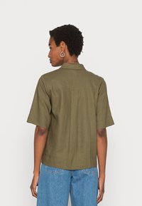 Camisa corta de mangas em verde-azeitona com colarinho e plissado nas costas, combinada com jeans azul-claro. O tecido tem uma aparência texturizada, semelhante ao linho.