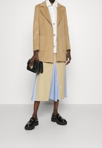 Person in einem beigen Mantel, weißem Hemd mit bunten Knöpfen, beige-blauem Rock, schwarzen Plateauschuhen und einer schwarzen Clutch.