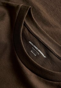 Pruun puuvillane T-särgi krae, millel on sile tekstuur ning valge silt, millel on brändi nimi, päritolu ja suuruse üksikasjad.