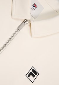 Camisa polo blanca con cuello y una cremallera plateada. Presenta un logo negro bordado en el lado izquierdo. Textura de tela suave.