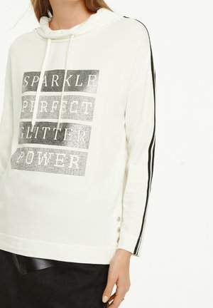 Femme portant un sweat à capuche blanc à manches longues avec des rayures noires sur les manches et un texte pailleté "SPARKLE PERFECT GLITTER POWER" sur le devant.