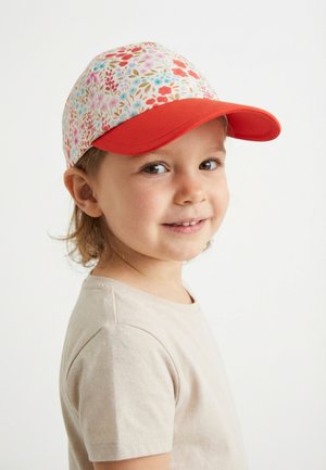 Junges Kind trägt ein beiges Hemd und eine Kappe mit Blumenmuster und rotem Schirm, lächelt sanft und steht leicht seitlich vor einem schlichten Hintergrund.