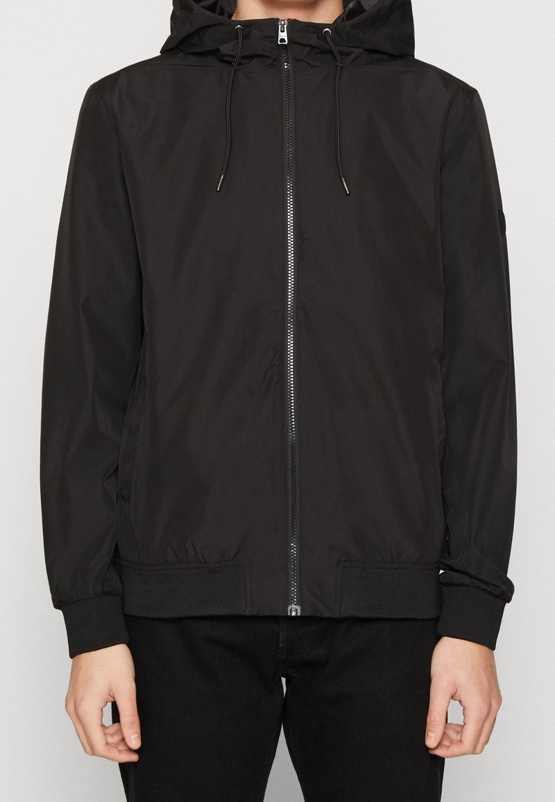 Personne portant une veste à capuche noire zippée avec cordons et poignets côtelés, debout devant un fond clair uni.