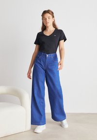 Femme portant un t-shirt noir col V, un jean bleu taille haute à jambes larges, et des baskets blanches, debout près d'un fauteuil blanc contre un mur uni.