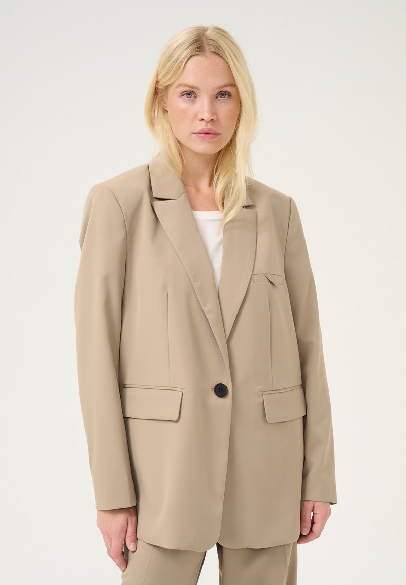 Blazer beige avec une coupe ajustée, un bouton noir unique, poches à rabat et col cranté, fabriqué à partir d'un tissu lisse et léger.