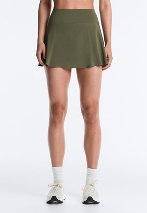 Žena v olivovo zelenej športovej sukni, bielych ponožkách a bielych bežeckých topánkach stojaca pred jednoduchým bielym pozadím.