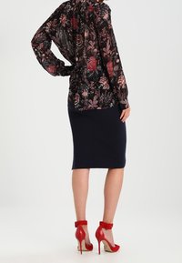 Blusa nera a maniche lunghe con fiori rossi e rosa, abbinata a una gonna a matita blu navy aderente e décolleté rossi con cinturini alla caviglia.