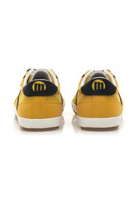 Zapatillas de ante amarillas con acentos negros, suelas blancas, cuello acolchado y detalle del logo en el talón. La vista trasera resalta el diseño y la costura.