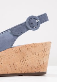 Sandale compensée en suédine bleue avec une plateforme en liège, dotée d'un détail de sangle à boucle ronde. La texture lisse et le motif naturel du liège mettent en valeur le design.