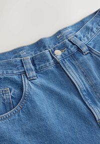 Gros plan sur la taille d'un jean ballon bleu avec bouton en métal, passants de ceinture, poche avant, et taille imprimée 34R sur la ceinture intérieure.