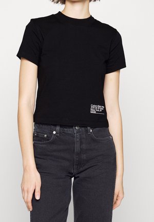 Osoba ubrana w dopasowany czarny krótki top z krótkim rękawem z małym białym napisem Calvin Klein Jeans w pobliżu brzegu oraz wysokowysokie czarne dżinsy.