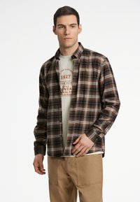 Braun- und beige-kariertes Hemd aus gewebtem Stoff, mit Knöpfen und Kragen. Darüber trägt er ein hellgrünes T-Shirt mit grafischem Druck.
