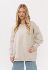 Przesadny beżowy sweter z okrągłym dekoltem, długimi rękawami i kieszenią z przodu; wykonany z miękkiego materiału z ribbowanymi mankietami i dołem.