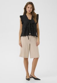 Blusa nera con dettagli in occhio di pernice e un nodo frontale, abbinata a pantaloni corti larghi beige. Scarpe nere con tacco sottile completano il look.