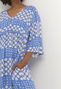 Blå och krämfärgad mönstrad klänning med V-ringning, korta ärmar, veck och fickor. Tillverkad av lätt tyg med en mjuk textur.