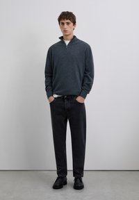 Joven de pie con las manos en los bolsillos, vistiendo un suéter gris oscuro con cremallera, jeans negros y zapatos negros, contra un fondo plano.