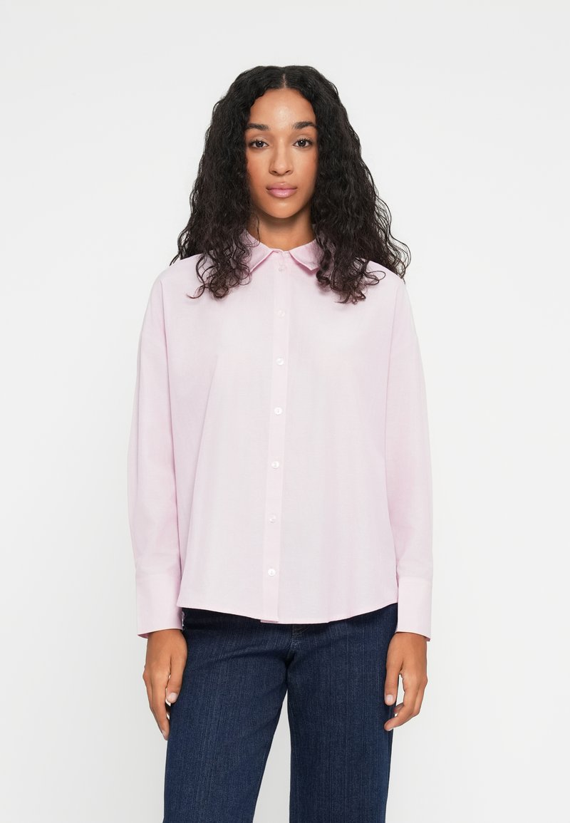 Chemise rose clair à boutons avec col, manches longues et coupe droite. Texture lisse, avec des boutons blancs sur le devant.