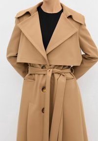 Personne portant un trench-coat beige ceinturé avec de grands revers et des boutons en écaille de tortue sur un haut noir, bras positionnés derrière le dos.