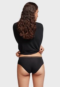 Zwarte katoenen slip met een scalloped tailleband, van achteren getoond. Draagt een bijpassend zwart losvallend shirt, waarmee het ontwerp van de slip wordt benadrukt.