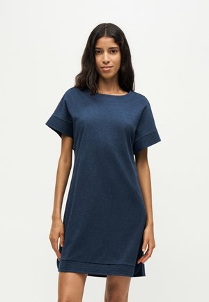 Frau mit langen dunklen Haaren, die ein locker sitzendes, kurzärmeliges dunkelblaues Kleid trägt und vor einem einfarbigen, hellen Hintergrund steht.