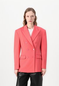 Dubbel-geknoopt koraalkleurig blazer gemaakt van gestructureerde stof, met schouderklappen, twee zakken en knoopdetails. Ggedragen over een wit top.