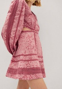 Femme portant une robe rose à motifs avec des manches bouffantes et un détail découpé à la taille, debout devant un fond clair uni.