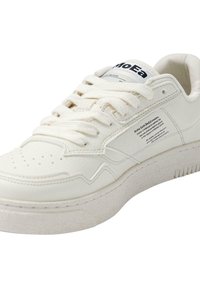 Hvite sneakers har en teksturert læroverdel, rund tå, tykk gummisåle og merking på tungen. Snøringene er flate og flettede.